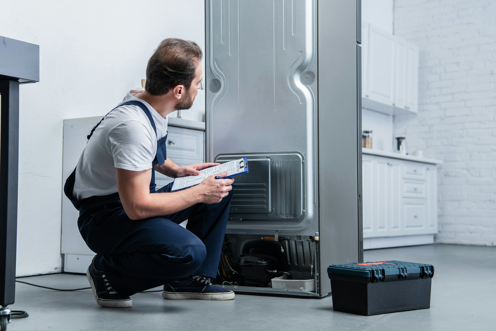 handyman in working overall writing in clipboard near broken refrigerator in kitchen